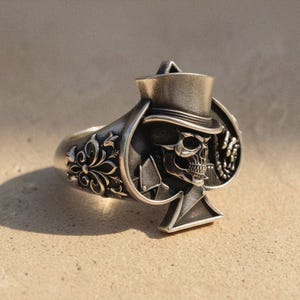May include: Silver ring with a skull in a top hat design, set within a spade shape. The ring band has decorative floral patterns. A distinctive piece of jewellery.