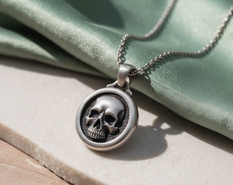May include: A silver-toned metal pendant necklace featuring a circular frame with a raised, detailed human skull design in the centre. The pendant is attached to a delicate silver-toned chain link necklace with a smooth, polished finish.