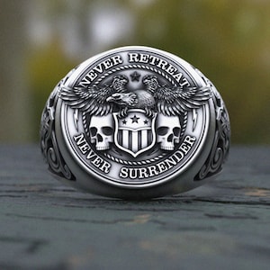 Pode incluir: Anel prateado com design detalhado. Apresenta uma águia com asas abertas, duas caveiras e um escudo com estrelas. As palavras "NEVER RETREAT" e "NEVER SURRENDER" estão inscritas ao redor do anel.