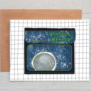 May include: A black and blue card with a starry sky and a moon inside. The card says "you're a keeper" in green text.