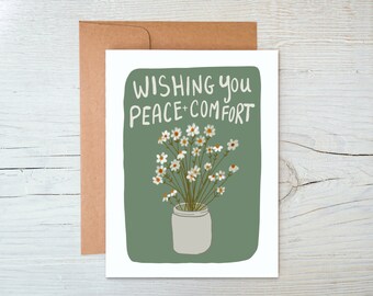 Puede incluir: Una tarjeta verde con margaritas blancas en un tarro blanco. La tarjeta dice "Wishing you peace + comfort".