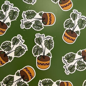 May include: Stickers featuring a potted Monstera plant with the text "Plant Daddy" in a handwritten font. The plant pot is brown with a tan and red geometric design. The background is a solid green.