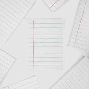May include: White notepads with light blue horizontal lines and a vertical red line on the left side. The notepads are arranged on a white surface. The notepads are blank and ready for writing.