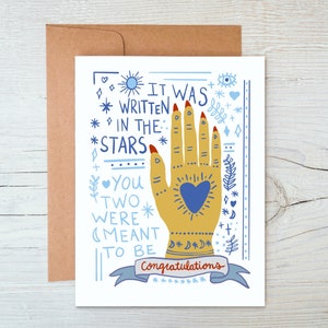 May include: A white card with a blue and yellow illustration of a hand with a heart in the palm. The text on the card reads "It was written in the stars. You two were meant to be. Congratulations."