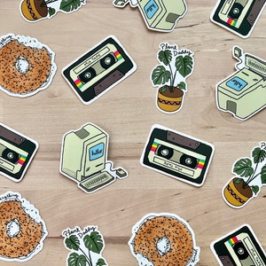 May include: A collection of stickers featuring a retro computer, cassette tapes, bagels, and potted plants. The stickers have a hand-drawn style with text that reads "hello", "MIX TAPE", "Plant Daddy", and "you're my everything". The color palette includes beige, brown, green, and black.