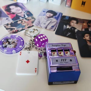 May include: A collection of fan merchandise featuring images of a musical group. Items include photo cards, a purple die, a slot machine, a keychain with a heart-shaped clasp, and a playing card.