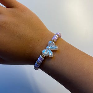 Puede incluir: Una pulsera de cuentas con un dije de lazo. La pulsera tiene cuentas de color lavanda, blanco y dorado. El dije de lazo es blanco nacarado. Diseñada para ser usada en la muñeca.