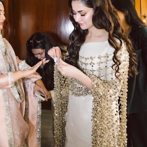 May include: A woman in a white dress with gold embellishments and a matching shawl. The shawl is made of gold sequins. Other women are in the background, wearing pink and black outfits.