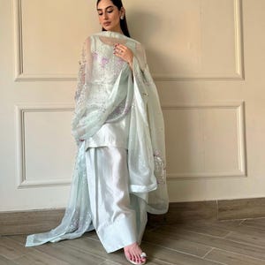 May include: A light blue, three-piece outfit featuring a tunic, wide-leg pants, and a sheer shawl. The shawl is adorned with sequins and floral embroidery. The outfit is made of a silky fabric and is suitable for formal occasions.