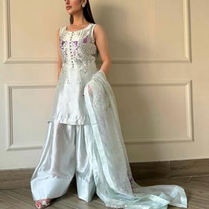 May include: A sleeveless, light blue outfit with wide-leg pants and a matching sheer scarf. The top is embellished with silver and purple sequins. The outfit is made of a shiny, silky material, and the scarf is draped over the arm.