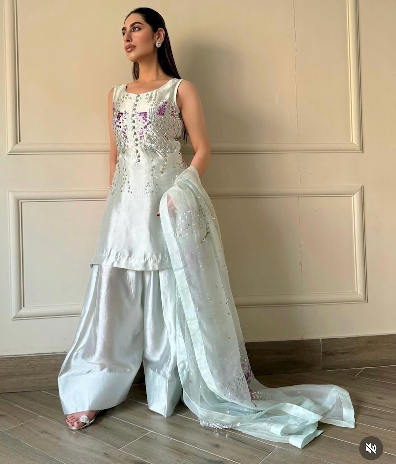 May include: A sleeveless, light blue outfit with wide-leg pants and a matching sheer scarf. The top is embellished with silver and purple sequins. The outfit is made of a shiny, silky material, and the scarf is draped over the arm.