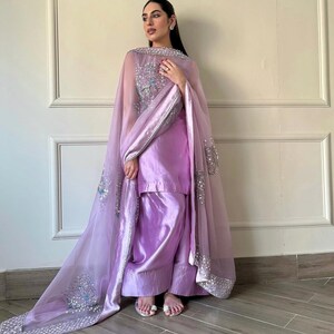 May include: A lilac-colored, traditional South Asian outfit. The ensemble includes a tunic, wide-leg pants, and a sheer, embellished shawl. The shawl features intricate silver and blue sequin detailing along the edges and across the shoulders. The outfit is made of a shiny, satin-like fabric.