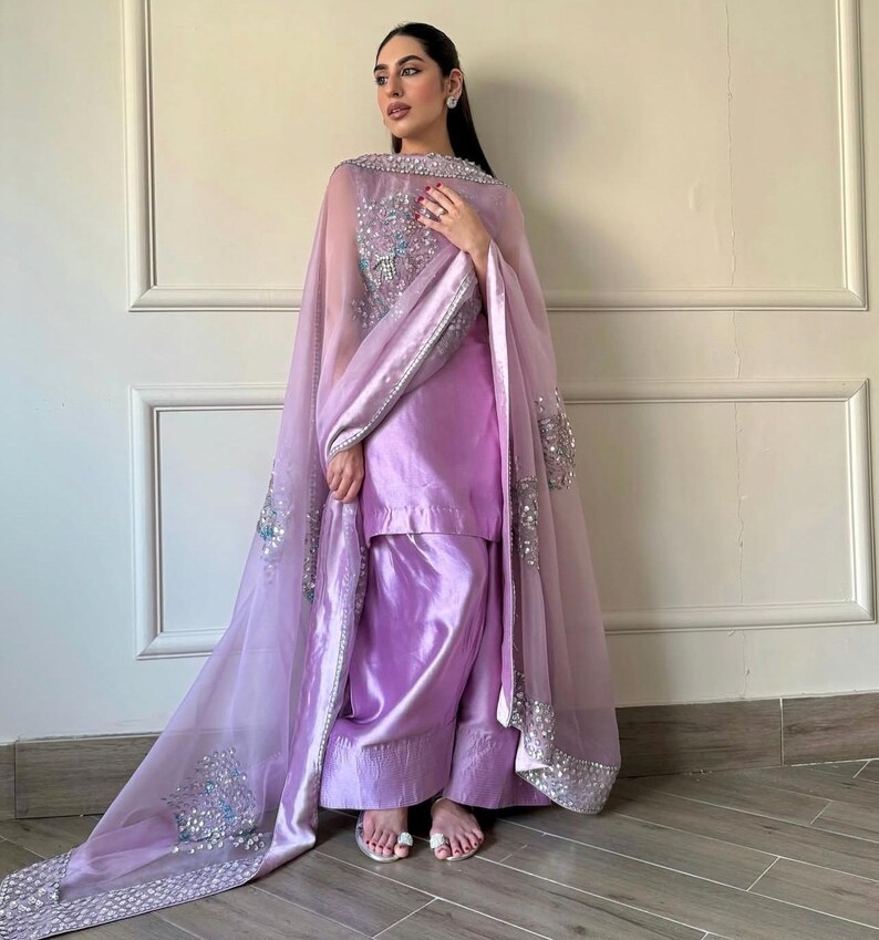 May include: A lilac-colored, traditional South Asian outfit. The ensemble includes a tunic, wide-leg pants, and a sheer, embellished shawl. The shawl features intricate silver and blue sequin detailing along the edges and across the shoulders. The outfit is made of a shiny, satin-like fabric.
