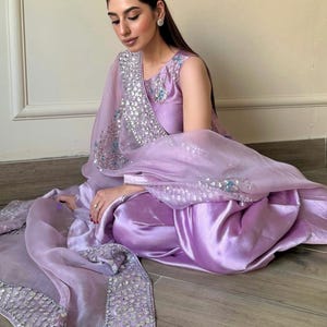 May include: A lilac-colored, sleeveless salwar kameez set. The outfit includes a satin-like top and pants, paired with a sheer dupatta. The dupatta and neckline are embellished with silver sequins and light blue accents. The ensemble is worn by a person.