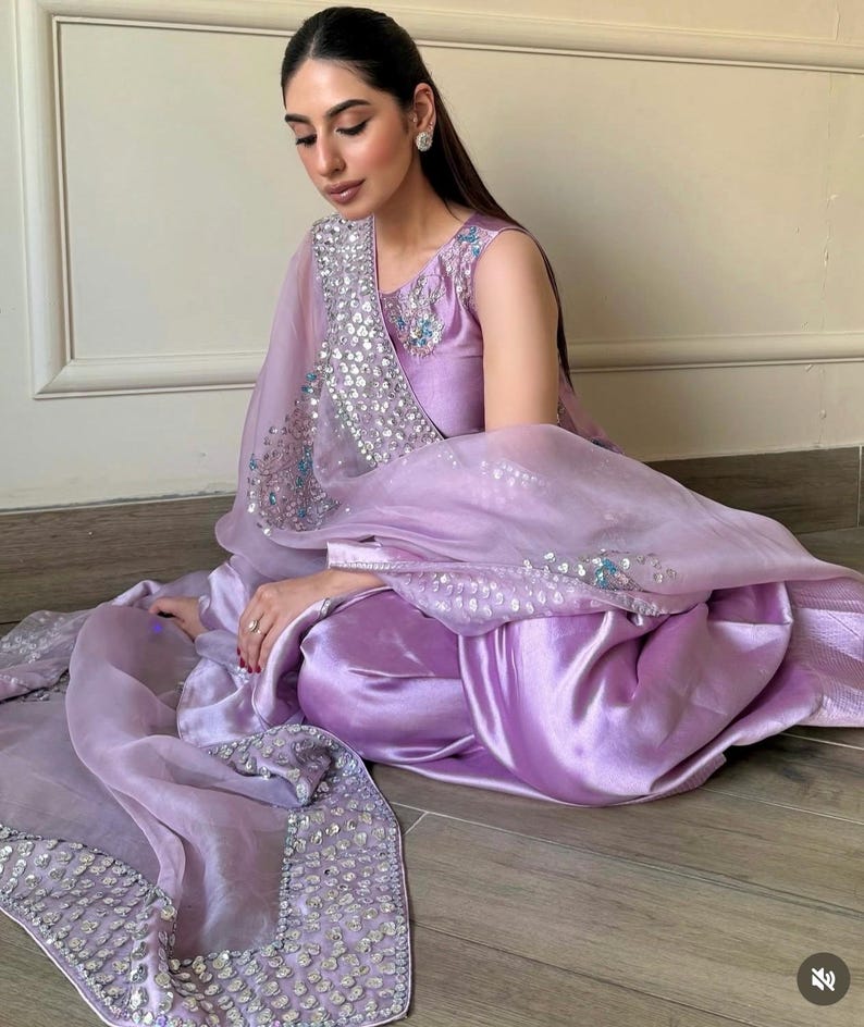 May include: A lilac-colored, sleeveless salwar kameez set. The outfit includes a satin-like top and pants, paired with a sheer dupatta. The dupatta and neckline are embellished with silver sequins and light blue accents. The ensemble is worn by a person.