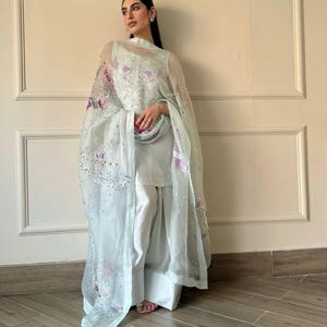 May include: A light blue ensemble featuring a sleeveless top, wide-leg pants, and a sheer, embellished shawl. The shawl is adorned with sequins and purple floral embroidery. The outfit is a soft, pastel color, creating a delicate and elegant look.