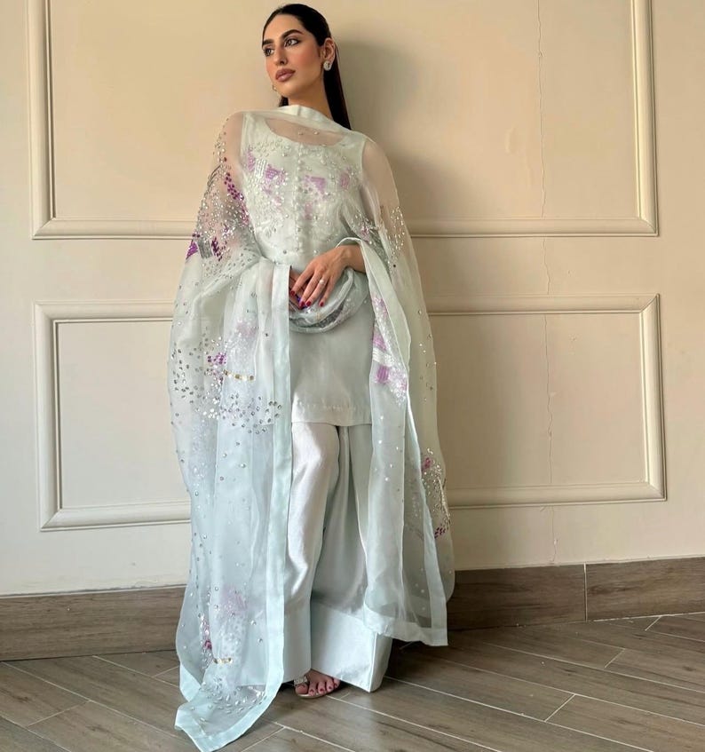 May include: A light blue ensemble featuring a sleeveless top, wide-leg pants, and a sheer, embellished shawl. The shawl is adorned with sequins and purple floral embroidery. The outfit is a soft, pastel color, creating a delicate and elegant look.