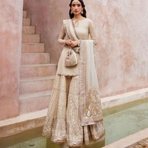 May include: An ivory-colored, long-sleeved dress with intricate embroidery and a matching purse. The dress features a tiered skirt and a sheer, embellished shawl. The outfit is accessorized with delicate jewelry, creating an elegant ensemble.