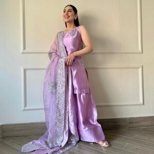 May include: A sleeveless, lilac-colored formal outfit. The ensemble includes a top, wide-leg pants, and a sheer scarf with silver embellishments. The outfit is made of a shiny fabric, and the scarf drapes elegantly.