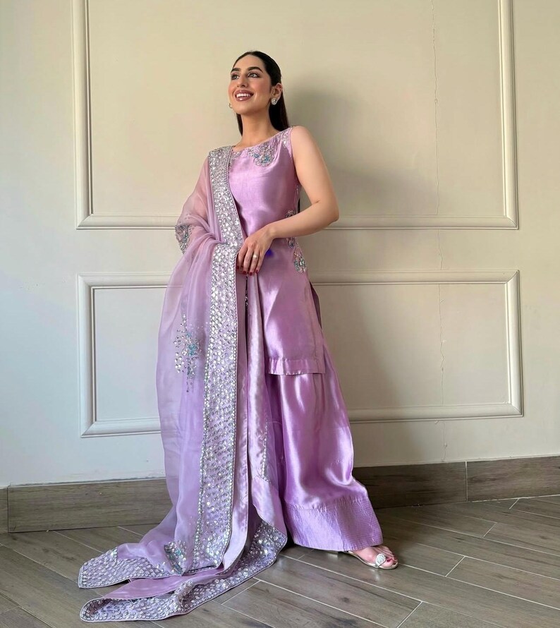 May include: A sleeveless, lilac-colored formal outfit. The ensemble includes a top, wide-leg pants, and a sheer scarf with silver embellishments. The outfit is made of a shiny fabric, and the scarf drapes elegantly.
