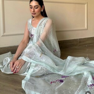 May include: A light blue formal outfit featuring a sleeveless top, matching pants, and a sheer scarf. The outfit is embellished with sequins and embroidery in shades of purple and silver. The ensemble is worn by a person sitting on a wooden floor.