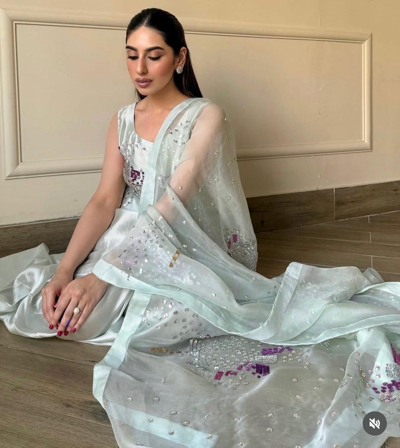 May include: A light blue formal outfit featuring a sleeveless top, matching pants, and a sheer scarf. The outfit is embellished with sequins and embroidery in shades of purple and silver. The ensemble is worn by a person sitting on a wooden floor.