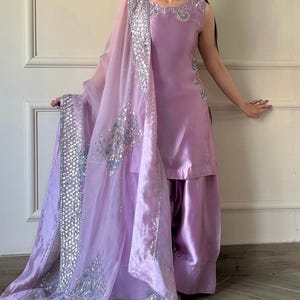 May include: A sleeveless lavender salwar kameez ensemble. The outfit includes a tunic top and wide-legged pants. A sheer dupatta with silver embellishments and embroidery drapes over the shoulders. The outfit is made of a shiny fabric.