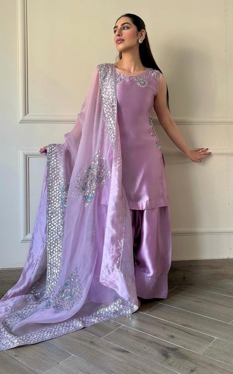 May include: A sleeveless lavender salwar kameez ensemble. The outfit includes a tunic top and wide-legged pants. A sheer dupatta with silver embellishments and embroidery drapes over the shoulders. The outfit is made of a shiny fabric.