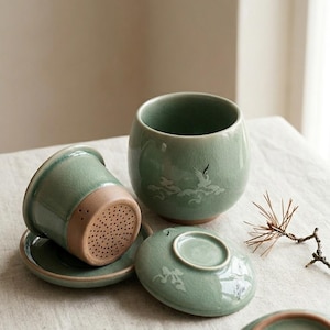 May include: A set of celadon green ceramic tea cups with matching saucers and lids. The cups have a rounded shape and feature a crane and wave design. The tea strainer is a light brown color. The set is displayed on a neutral surface.