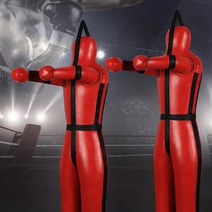 May include: Two red boxing training dummies with black straps. The dummies are standing in a boxing ring.