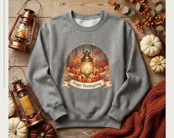 May include: A heather gray crewneck sweatshirt featuring a central graphic of a lit lantern surrounded by orange pumpkins and autumn leaves, with a banner below the lantern displaying the text Happy Thanksgiving in a classic serif font.