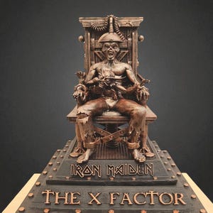 Op de afbeelding: Een bronzen sculptuur van een figuur vastgebonden aan een elektrische stoel. De sculptuur staat op een voetstuk met de woorden "IRON MAIDEN" en "THE X FACTOR" in bronskleurige letters. De figuur heeft een verontruste uitstraling.