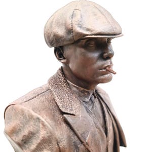 Peut inclure: Buste en bronze d'un homme portant une casquette de garçon de journal et une veste de costume, avec une cigarette à la bouche. La sculpture a une texture détaillée et est sur fond blanc.