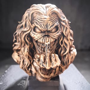 Puede incluir: Un busto de color bronce de una figura humanoide con cabello largo y ondulado, ojos hundidos y una amplia sonrisa con dientes. La figura sostiene una mano frente a su rostro, con el dedo medio extendido verticalmente en un gesto, montado sobre una base negra.