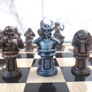 Op de afbeelding: Een schaakset met drie schaakstukken op een houten bord. De stukken zijn gemaakt van metaal en hebben een gedetailleerd ontwerp. Het stuk in het midden is een zilveren samurai-krijger met een helm en harnas. Het stuk links is een bruine krijger met een helm en harnas. Het stuk rechts is een bruine farao met een hoofdtooi.