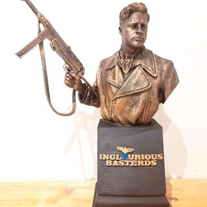 Puede incluir: Busto de color bronce de un hombre sosteniendo una ametralladora. El busto está montado sobre un pedestal negro con las palabras "INGLOURIOUS BASTERDS" y un logotipo. La escultura tiene un acabado detallado y está sobre un fondo claro.