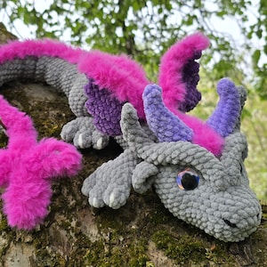 Pode incluir: Um brinquedo de dragão de crochê feito à mão em tons de cinza, roxo e rosa. O dragão tem olhos grandes e expressivos e está posicionado em um galho de árvore. O corpo do dragão é cinza, com detalhes em rosa e roxo.