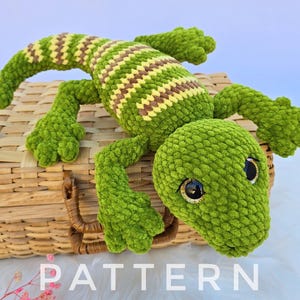 以下が含まれることがあります： 緑と茶色のストライプの背中を持つ手作りの緑色の鉤針編みのトカゲが、柳のバスケットの上に置かれています。トカゲは大きな黒い目を持ち、柔らかい糸で作られています。「PATTERN」という文字が白い文字で表示されています。