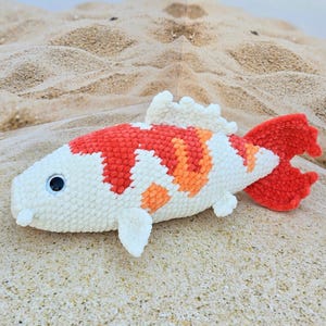 Pu&ograve; includere: Un giocattolo di pesce koi all'uncinetto con colori bianco, arancione e rosso. Il pesce &egrave; sdraiato su una superficie sabbiosa.