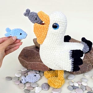 May include: A white, black, and yellow plush pelican with a fish in its beak, sitting on a rock. A hand holds a blue plush fish, and there are small pebbles scattered around the base. The pelican has blue eyes.