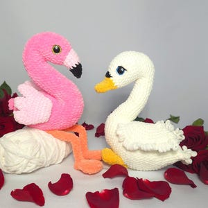 Puede incluir: Un juguete de peluche rosa en forma de flamenco y un juguete de peluche blanco en forma de cisne. Ambos juguetes están hechos a crochet y tienen ojos grandes y negros. El flamenco está sentado sobre una bola de hilo blanco y el cisne está sentado sobre un lecho de pétalos de rosa rojos.