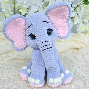 May include: A crocheted elephant toy in shades of blue and pink. The elephant has large, pink-lined ears, blue eyes, and black stitched details. It is sitting on a cream-coloured, lacy surface, with white flowers in the background.