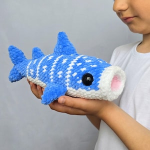 May include: A blue and white crocheted whale shark plush toy with a white belly and pink mouth. The plush toy has black button eyes and is held in a person's hands. The toy is a soft, handmade item.