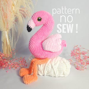 May include: Pink crocheted flamingo toy with orange legs. The flamingo is sitting on a white yarn ball. The text "pattern no sew!" is written in black on a white background.