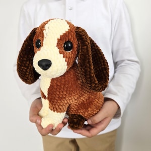 May include: A handmade plush dog toy with brown, cream, and white colors. The dog has large black eyes, long floppy ears, and a small tail. The toy is made of soft, textured material.