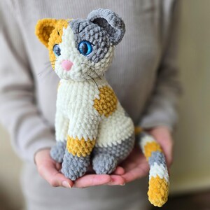 May include: A handmade plush cat toy in white, gray, and yellow. The cat has blue eyes, a pink nose, and black whiskers. The cat is being held in someone's hands.
