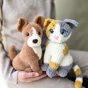 Może przedstawiać: Dwa ręcznie robione szydełkowe pluszaki: brązowy pies z białymi znaczeniami i kot szylkretowy. Pies ma białą plamę na pyszczku i czarny nos. Kot ma niebieskie oczy i różowy nos. Oboje siedzą prosto.