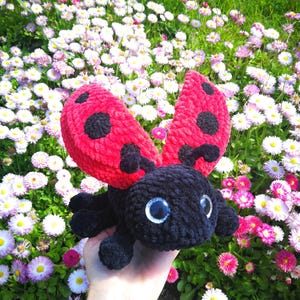 May include: A crocheted ladybug toy with black fur and red wings with black spots. The ladybug is being held in a hand against a background of white and pink flowers.