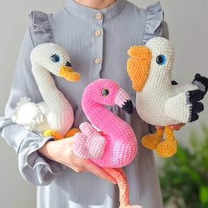 May include: Three handmade crochet bird plushies: a white swan with a yellow beak, a pink flamingo, and a white seagull with a black wing. Each plush has blue eyes and is held by a person wearing a gray dress. The flamingo has a long neck and legs.