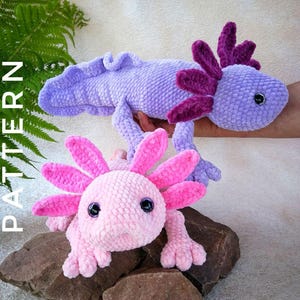 May include: Two crocheted axolotls in the image. One is a light pink color with dark pink gills and the other is a light purple color with dark purple gills. The axolotls are on a rock. The word "PATTERN" is printed on the left side of the image.